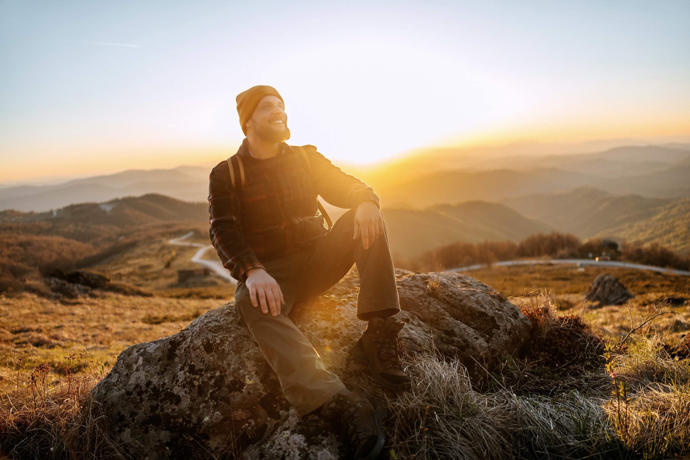 A man sitting on a rocky outcrop at sunrise, looking out over a mountain valley with contentment